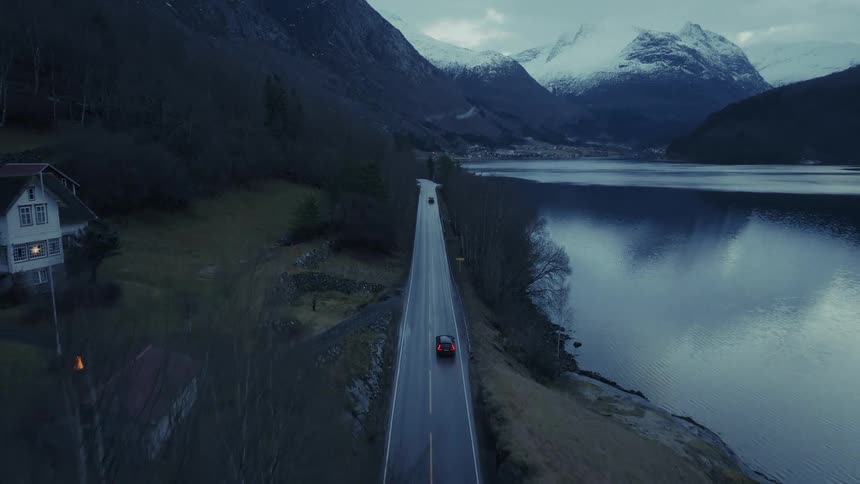 An aerial view shows a car driving on a road between a dark lake and a steep hill under a gray sky