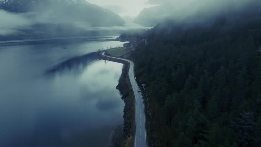 An aerial shot shows a single red car traveling along a winding road that hugs the edge of a calm misty lake densely forested mountains and lowhanging clouds create a cold atmospheric landscape surrounding the water