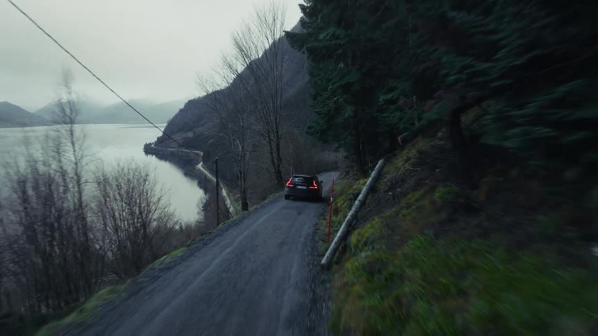 A black car drives along a narrow gravel road on a mountain side next to a large body of water under an overcast sky with pine trees and bare branches lining the path