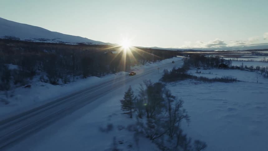 Automobiles traveling on a snow covered freeway through woodland as the sun shines over a mountain