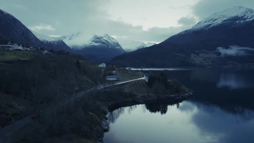 A dark car drives along a winding coastal road next to a calm body of water with snow covered mountains and a cloudy sky in the background during twilight
