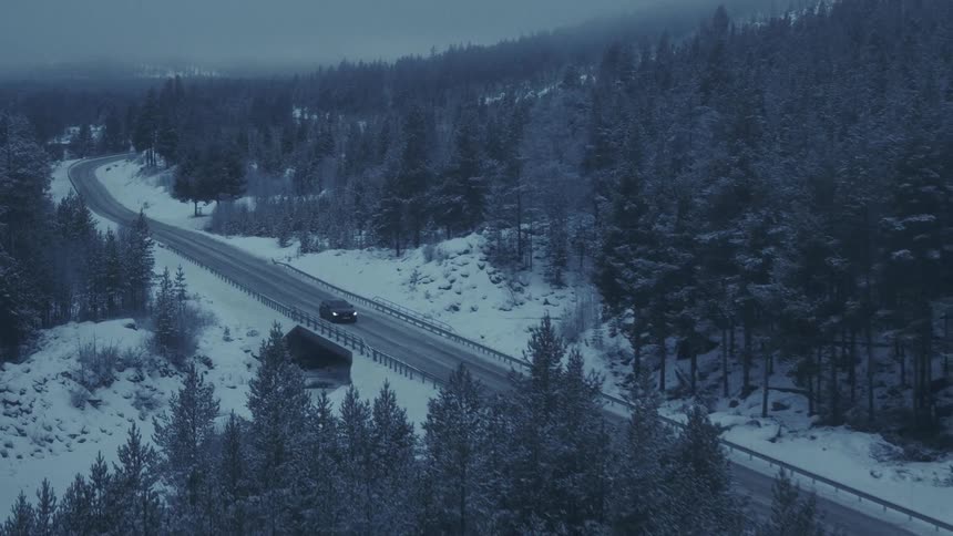 An aerial view shows a car as it drives on a road through a vast forest covered in deep snow and crosses over a small bridge during the evening