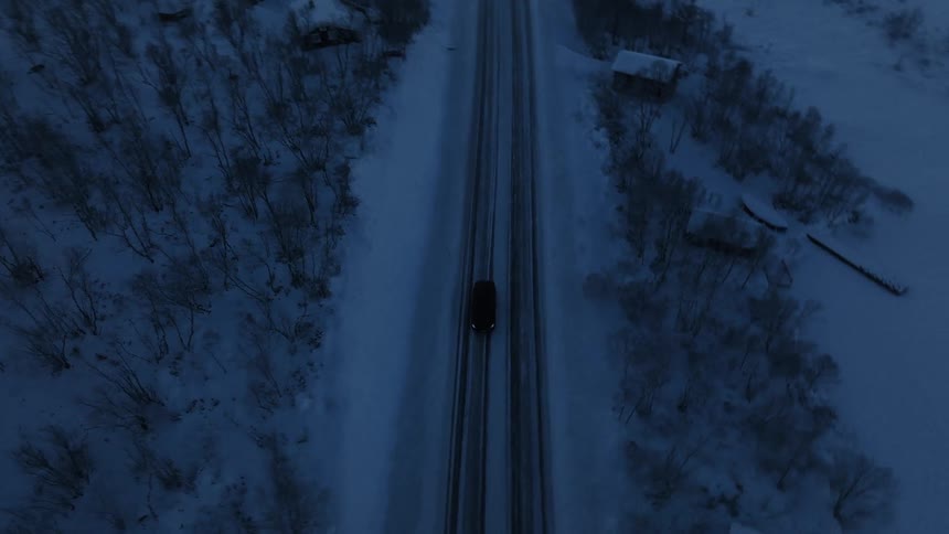 An aerial shot shows a dark car traveling along a snowcovered road that winds through a mountainous wintry landscapethe sun shines brightly over the horizon casting a cold glow across the scattered cabins and dense frostcovered trees lining the r