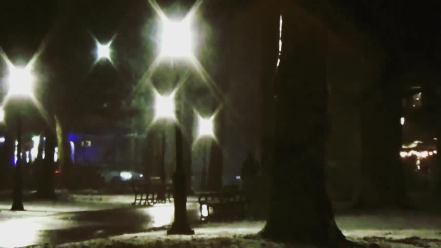 The video captures a dimly lit snowy park at night with glowing streetlights casting a hazy reflection on the ground shadows of trees and park benches create a quiet atmospheric scene as soft light flickers in the distance