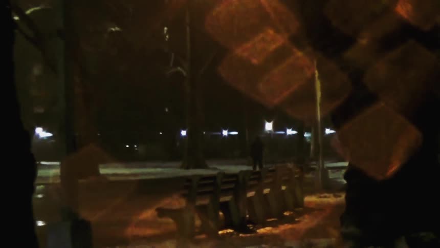 The video captures a dimly lit snowy park at night featuring empty benches and silhouettes of trees against distant streetlights large ambercolored bokeh or lens flares drift across the frame creating a dreamy and somewhat mysterious atmosphere