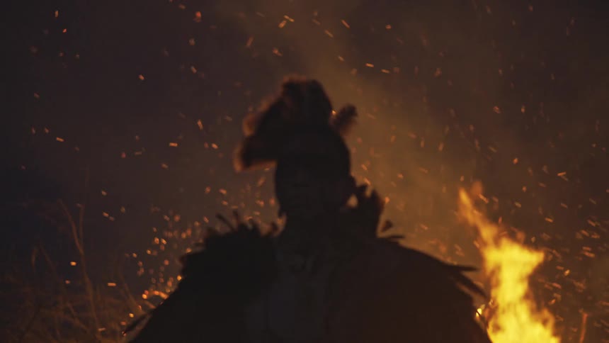 A person in a feathered costume stands as a silhouette in front of a large fire with orange sparks and flames rising into the dark night sky