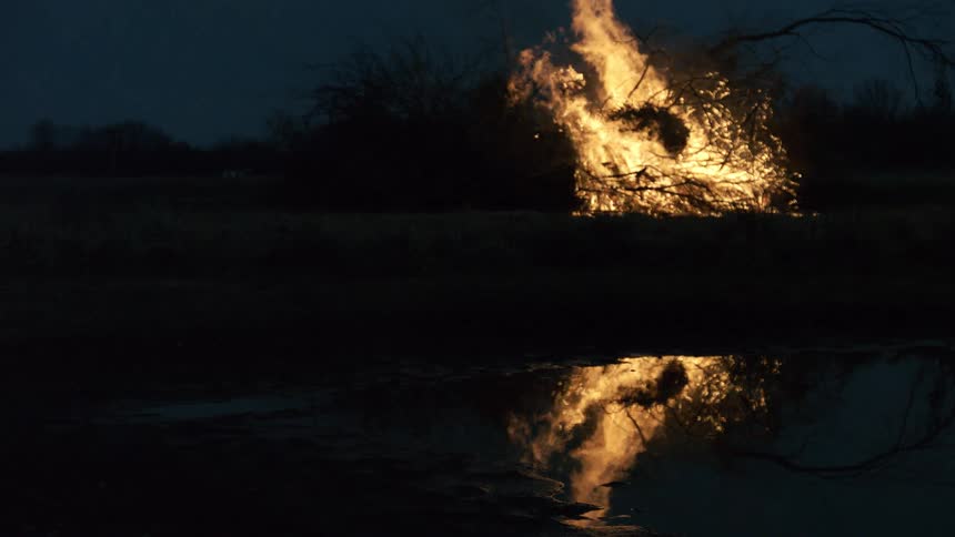 A large bonfire of dry branches blazes intensely in a dark open field at dusk the vibrant orange flames and glowing sparks are mirrored clearly in a still pond in the foreground