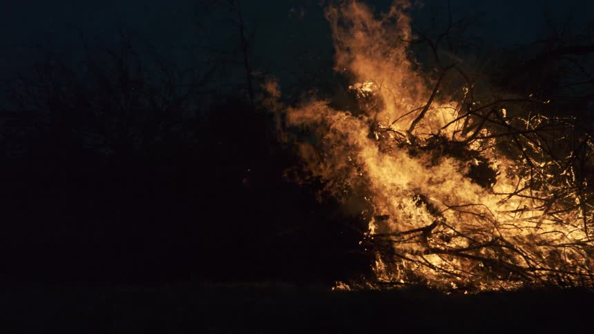 A brush fire is consuming a pile of branches with flames in slow motion creating a fluid flame effect as it throws smoke into a blackened night sky