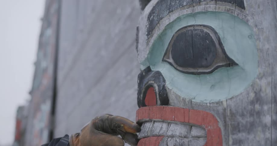 A person wearing a rugged tan work glove gently touches the carved wooden teeth of a traditional totem pole the camera slowly tilts upward to reveal the intricate painted details of the figures face against a neutral overcast background