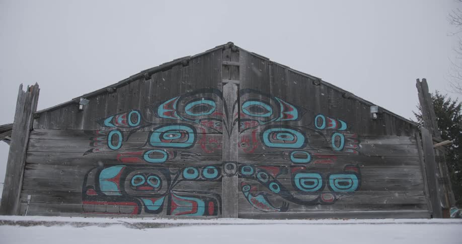 A wide view shows a large wooden building with a colorful mural of indigenous art on its front wall during a snowfall
