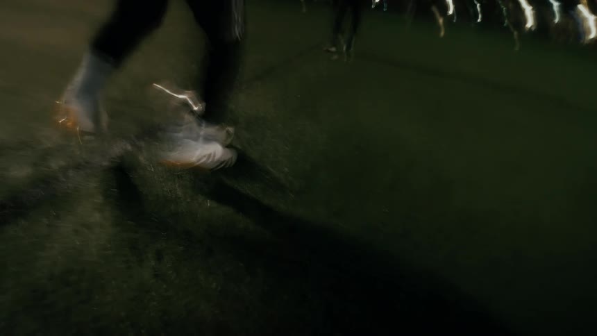 A motionblurred closeup shows a player in white cleats sprinting across a lit soccer field at night