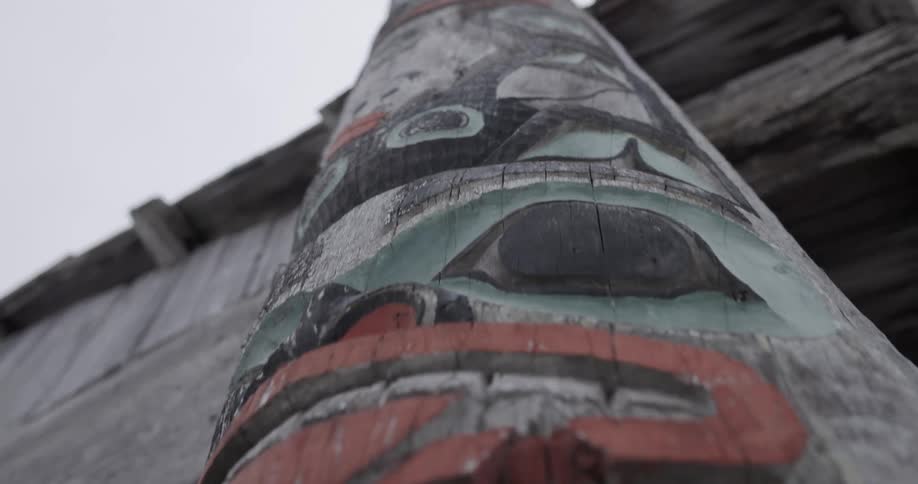 A low view moves upward along a weathered wooden totem pole featuring carved faces painted with red and green details against a grey sky