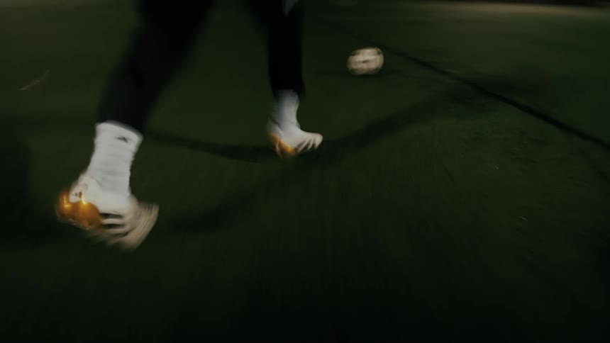 A player wearing white and gold cleats prepares to take a shot on an outdoor soccer pitch at nightthe camera follows the players feet as they move toward the ball with a goal and goalie visible in the background