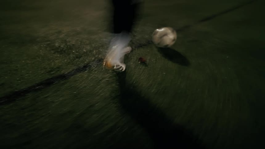 A close view of the feet of a man wearing white socks and soccer cleats as he dribbles a ball across a green turf field at night under bright stadium lights