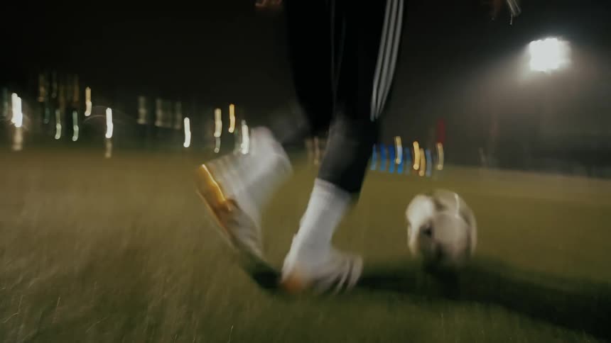 A closeup motionblurred shot shows a person in white cleats dribbling a soccer ball across a lit field at night while others on the field run along with the player
