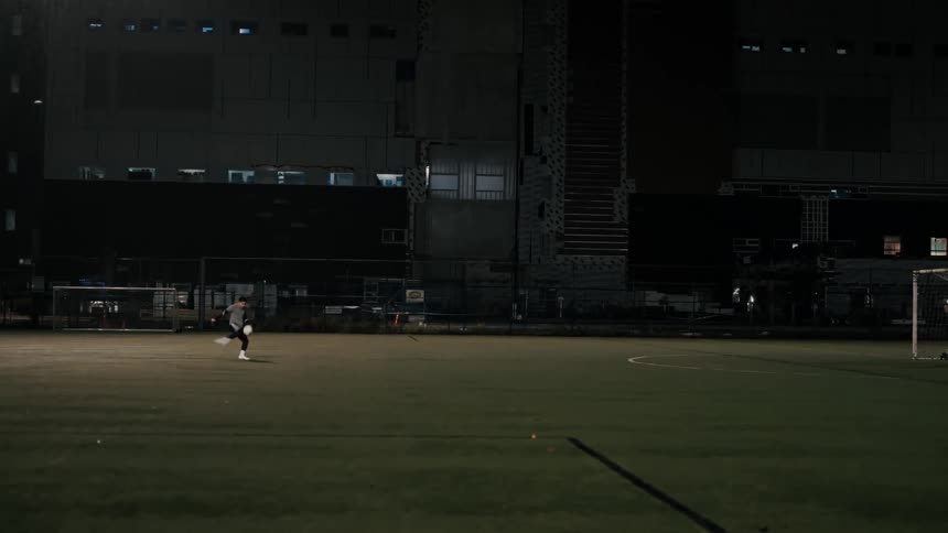 A distant view shows a soccer player kicking a ball across a green field at night toward a goal where a goalie stands ready to block the shot