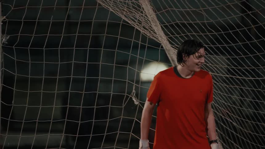 A young man with wet hair wearing an orange shirt and goalie gloves standing in front of a soccer net he is smiling and laughing while looking offcamera during a nighttime game or practice