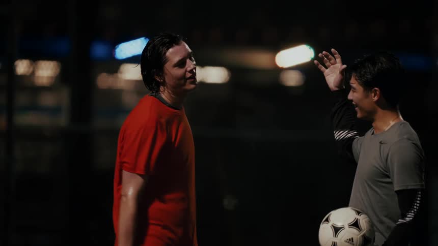 Two men wearing athletic clothes stand on a soccer field at night and celebrate by bumping fists and hugging