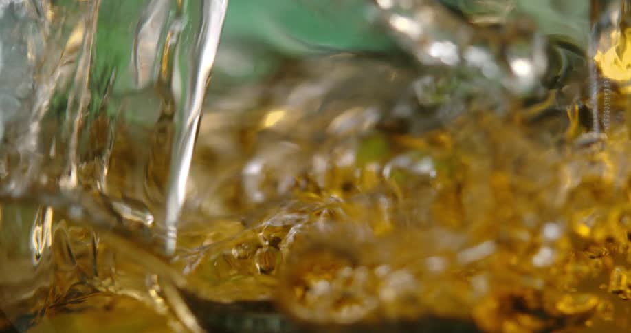 An amber liquid pours into a glass in extreme slow motion creating a chaotic splash and a flurry of golden bubbles the turbulent surface eventually settles into a steady effervescent flow against a soft green background