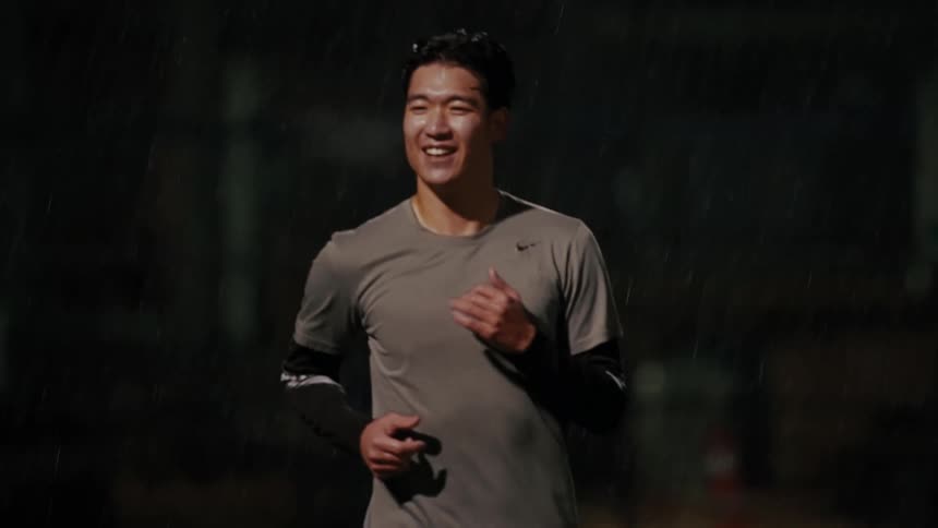 A smiling man wearing a grey athletic shirt and black leggings runs across a field in the rain at night under bright stadium lights while a blurred ball flies past in the background