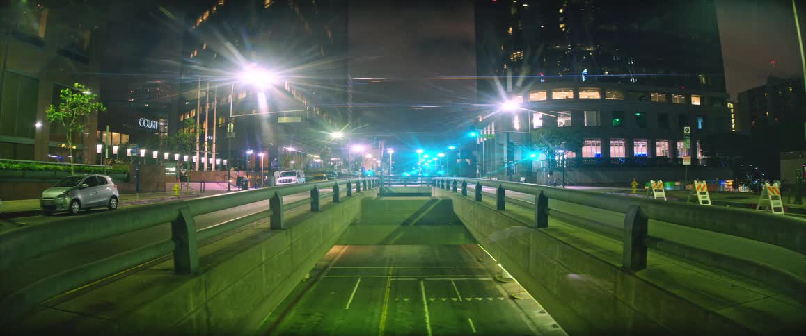 A brightly lit city street at night stretches over a concrete underpass with tall office buildings and glowing streetlights lining the scene