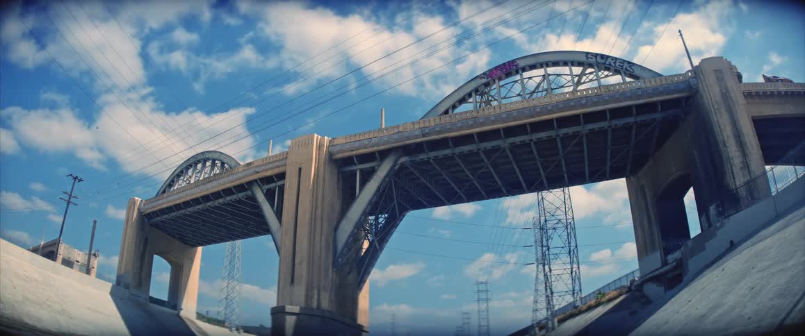 A large arched concrete bridge stretches across the frame under a bright blue sky with light traffic visible on the left and a small group of people gathered near the lower right along the sloped concrete embankment