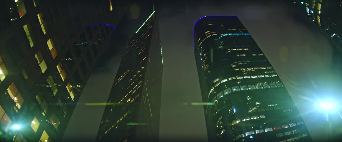 Looking up from a low angle massive illuminated skyscrapers tower into a dark cloudy night sky while subtle light leaks and reflections shimmer across the glass surfaces