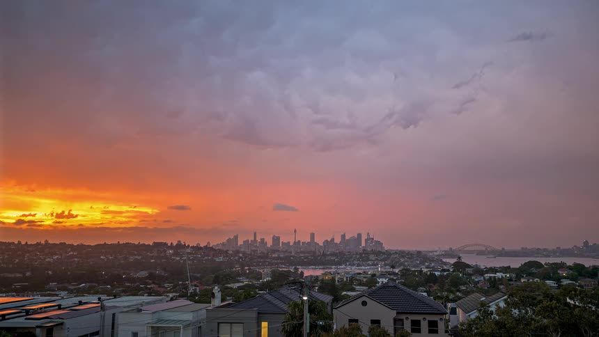 A wide view of a city skyline at dusk shows dark storm clouds overhead and a glowing orange sunset on the horizon beyond the rooftops in the foreground