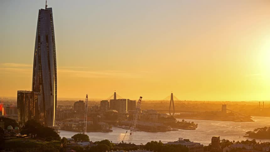 A time lapse shows the sun setting over a city harbor with a tall glass skyscraper and large construction cranes near the water as the sky turns from orange to dark blue