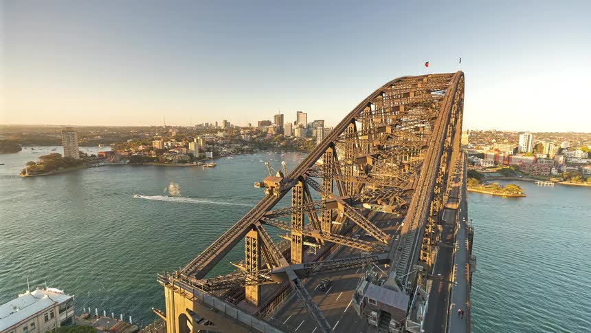 A busy bridge connecting two cities with cars driving across and boats passing underneath shown from midday through sunset into nighttime with two flags flying at the top of the bridge