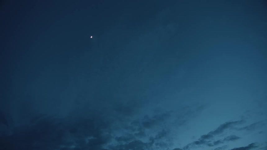 A slender crescent moon hangs high in a deep blue twilight sky as wispy dark clouds drift slowly across the lower frame creating a vast and tranquil nocturnal atmosphere