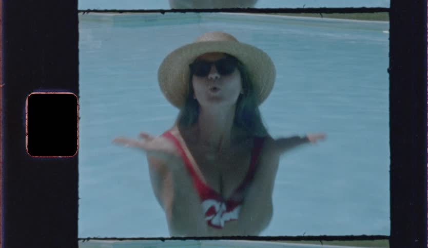 A woman wearing a red swimsuit and a straw hat with sunglasses blows a kiss while standing in a blue swimming pool in a vintage film style