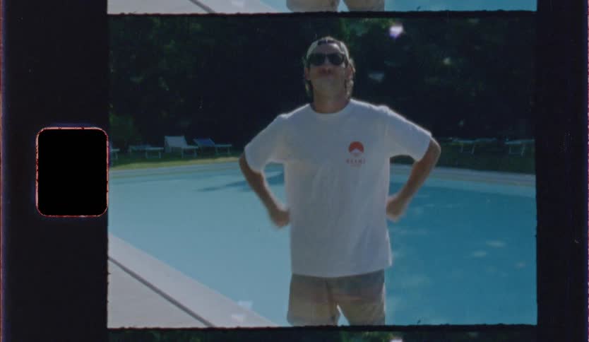 Vintage film footage shows a man wearing a white shirt and sunglasses standing in front of a blue swimming pool while putting on his sunglasses and smiling