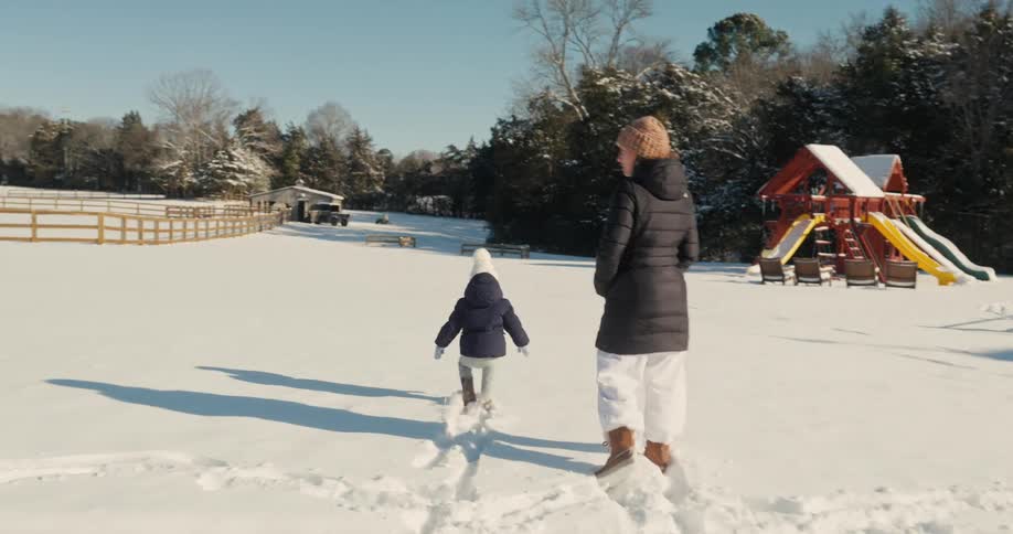 Adult and child walk on snow in a park with playground equipment in the background