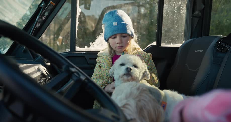 A young girl wearing a blue knit hat and a yellow patterned jacket sits in the driver seat of a vehicle while petting and hugging two small white dogs on a sunny day