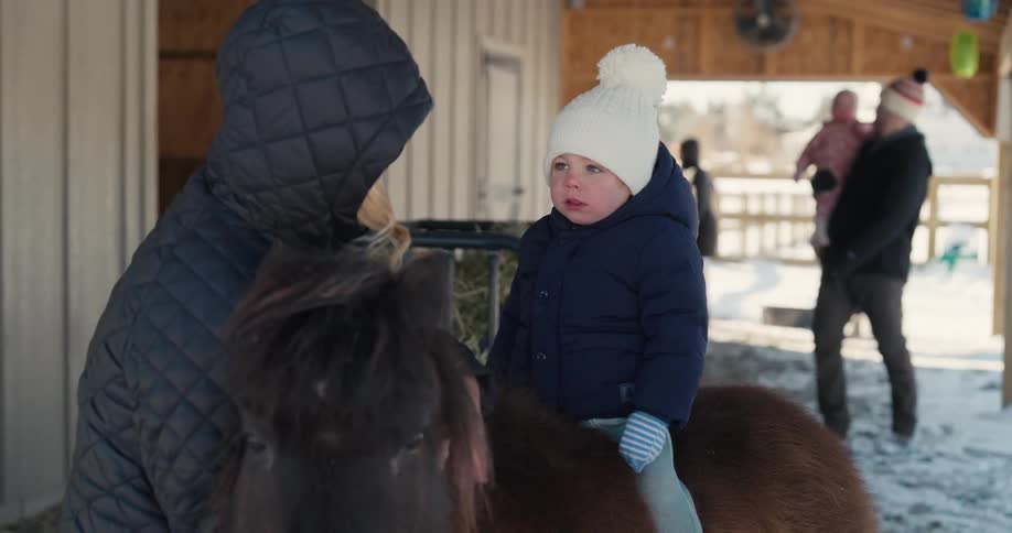 A toddler wearing a white knit hat and a dark blue coat sits on a brown pony while a woman in a black jacket stands beside them and a man carrying another child walks in the background at a farm