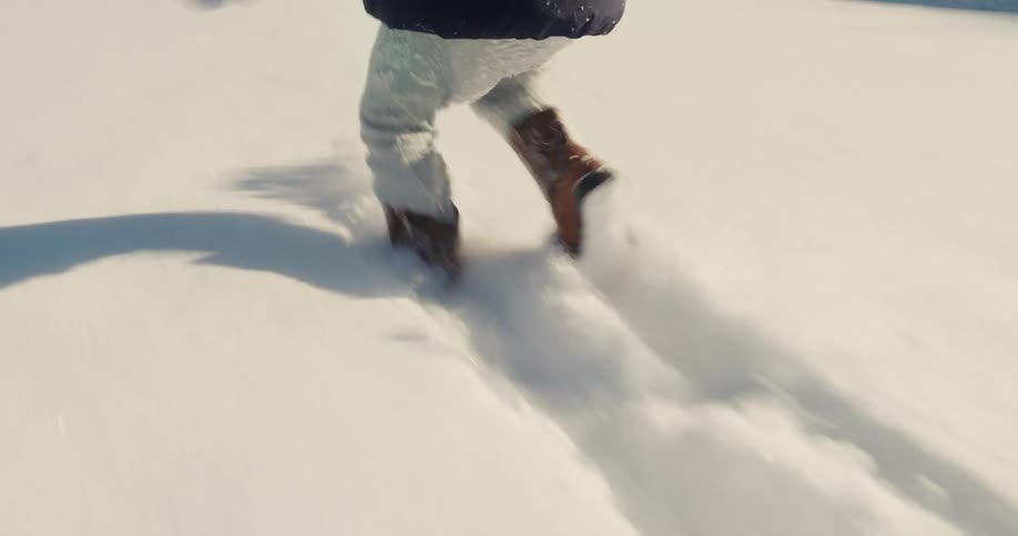 A young child in a white knit hat and navy jacket joyfully runs through a sunlit snowcovered yard the camera follows their legs closely as they kick up powdery snow while heading toward a wooden fence