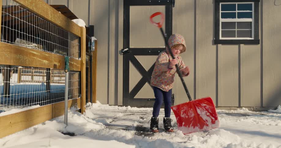 A young girl wearing a winter hoodie shoveling and packing snow outside a building at midday