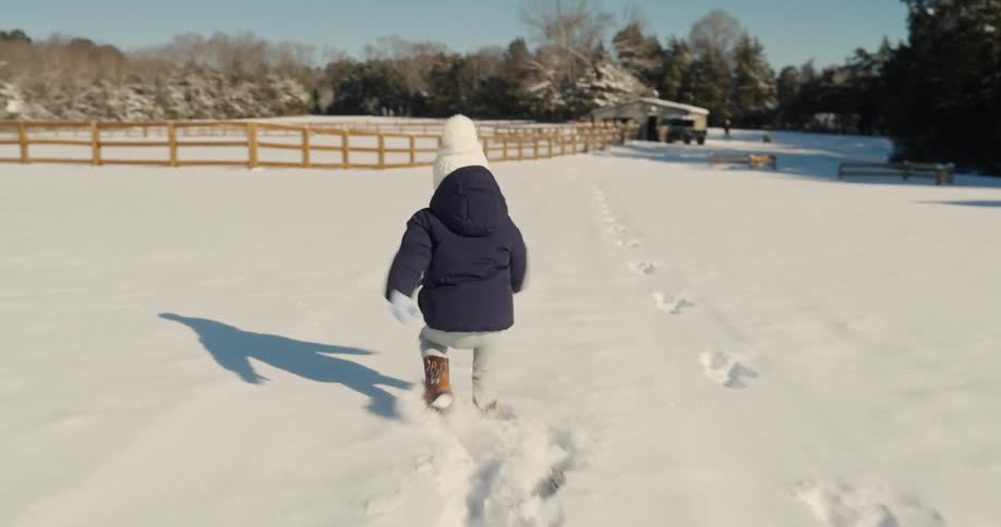 Child walks in the snow in an open field surrounded by a wooden fence