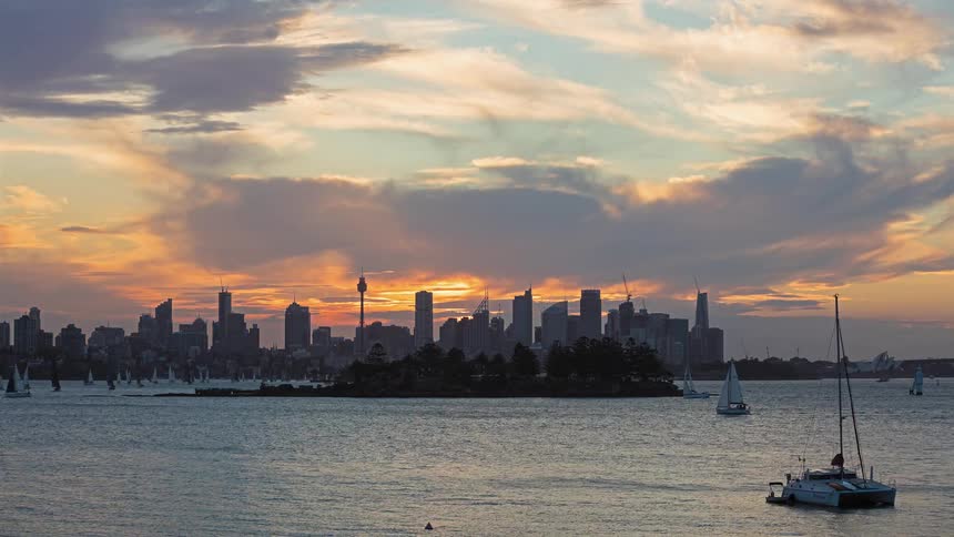 A time lapse view shows a golden sunset over a harbor with many sailboats on the water and a city skyline in the background