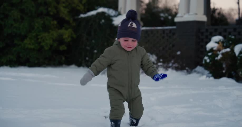 A young boy wearing a green snowsuit and a blue hat walks through deep white snow in a yard