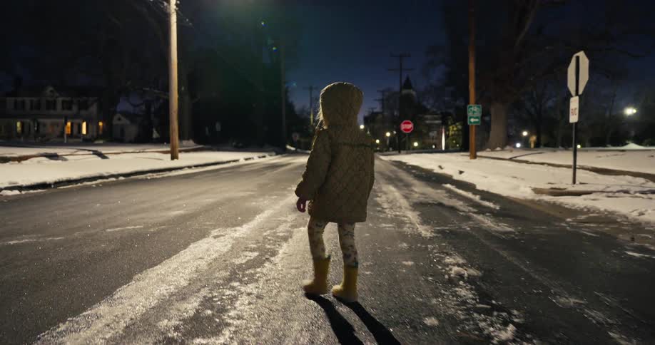 A small child wearing a hooded coat and yellow boots walks slowly down the center of a dark suburban street covered in patches of ice and snow during a cold winter night