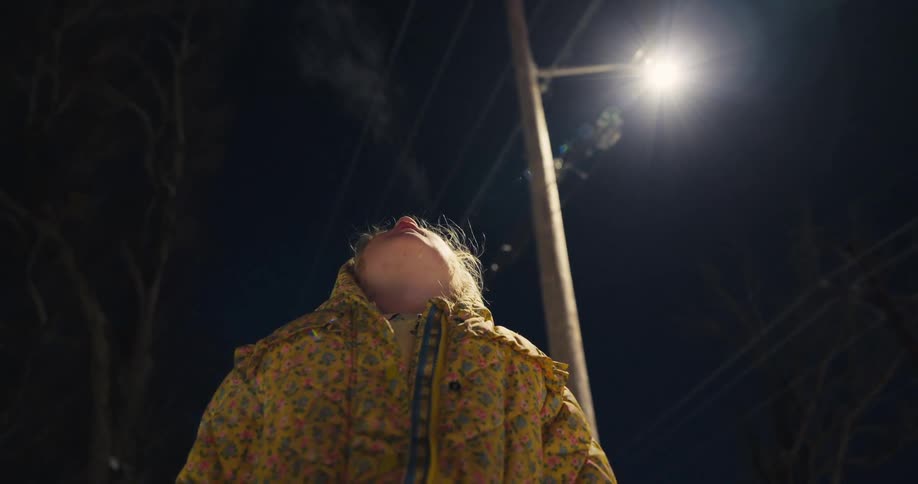 Person wearing a patterned jacket looking upward under a lit streetlight at night