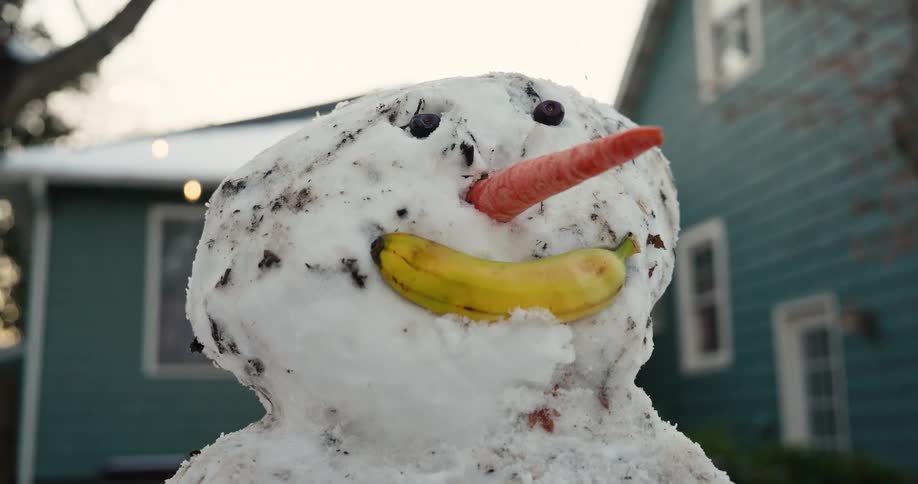 A close up shot of a snowman head featuring a large carrot for a nose and a yellow banana used as a smiling mouth while a blue house with string lights sits blurred in the background on a cold winter day