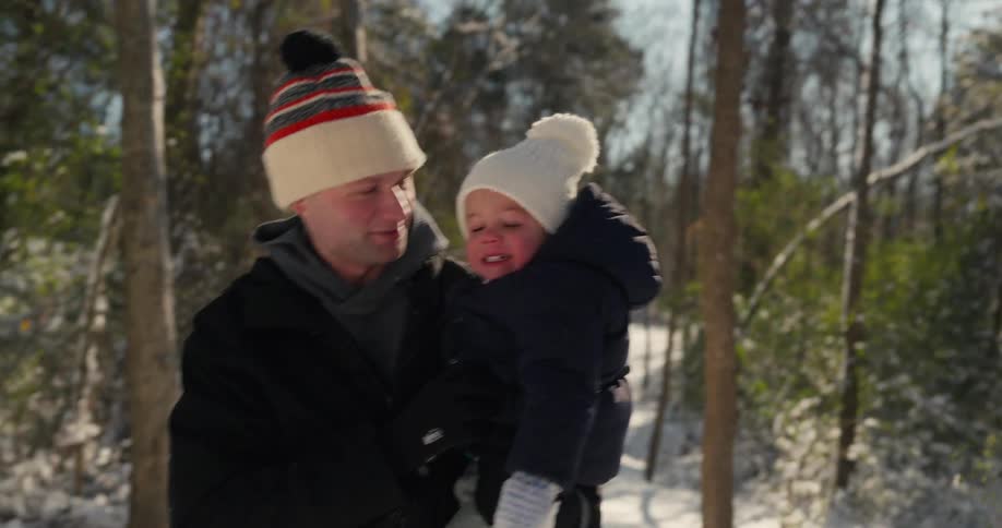 A man wearing a black jacket and a knit hat carries a young child in a coat and white hat while walking through a sunny forest covered in white snow