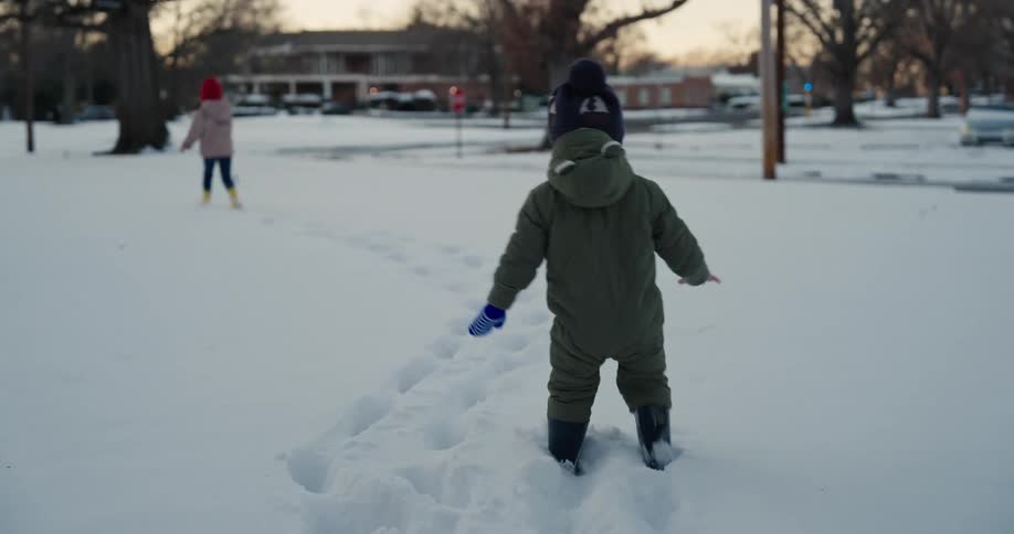 A small child in a green snowsuit and blue mittens walks through deep snow toward two other children in a neighborhood park during a winter evening