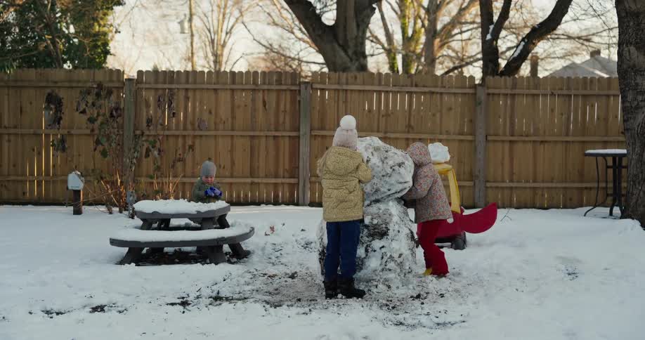 Children building a snowman in a fenced backyard with a picnic table nearby