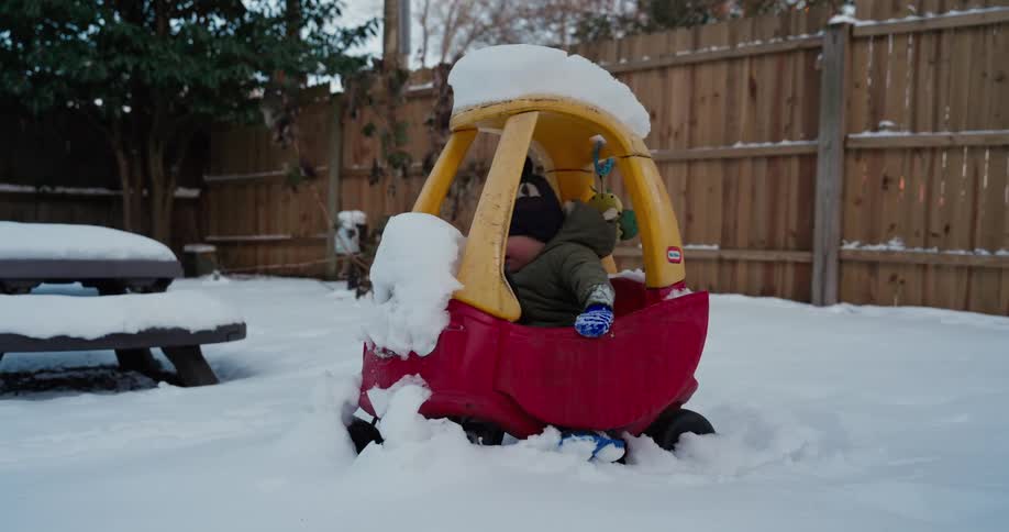 A child wearing a beanie and winter clothing gets into a toy car in the snow in the backyard of a house with a wooden fence at midday