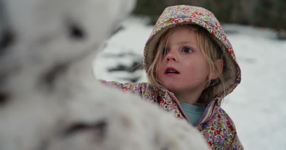 Little girl wearing a floral coat and hat touching a snowman in a snowy setting