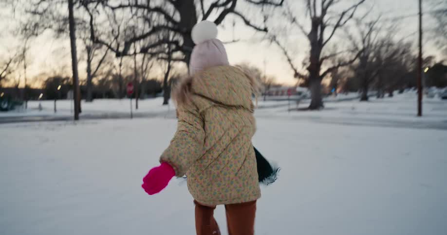 A young girl wearing a beige jacket and pink mittens runs excitedly through a snow covered park toward a large tree during a cold winter sunset while residential buildings and quiet streets are visible in the distance